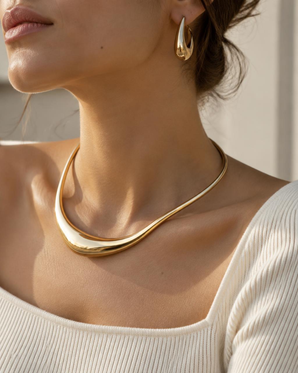 Close view of a woman wearing a sculptural gold necklace and gold earring with an ivory knit.